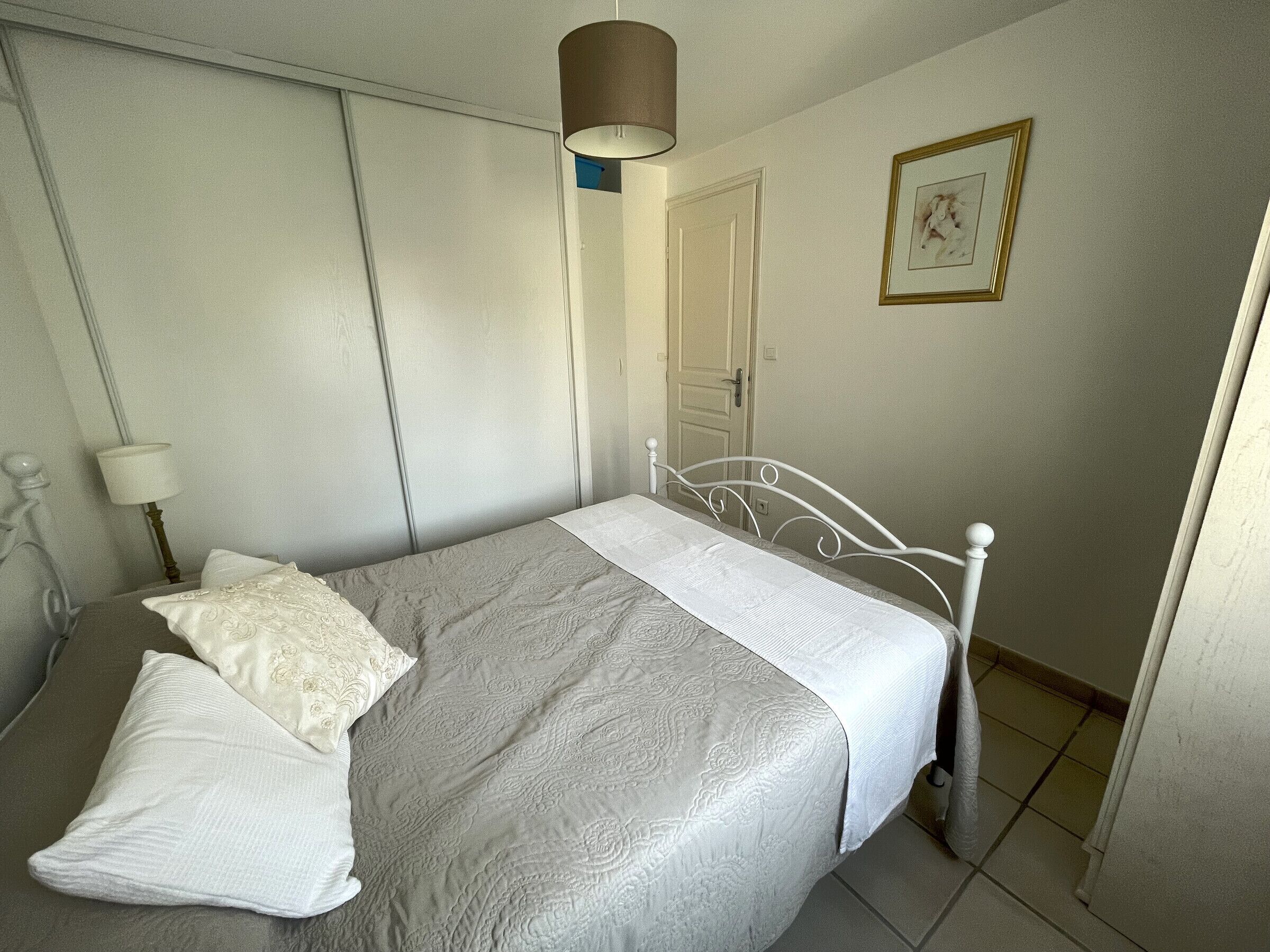 Una cama bien hecha con colcha y almohadas estampadas, cabecera de metal blanco, lámpara de noche y un cuadro en la pared.
