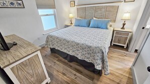 Une chambre à coucher avec un grand lit, deux tables de chevet avec des lampes, une fenêtre avec des stores et un tableau encadré au mur.