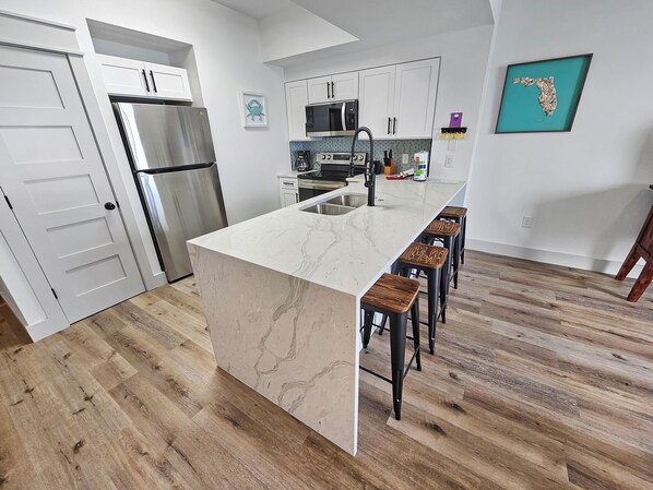 A modern kitchen with a marble island, stainless steel appliances, and wooden bar stools. - Ariel Dunes 2005 | Gulf Views from 20th Floor | Fully Renovated | Zero-Entry Pool (Miramar Beach)