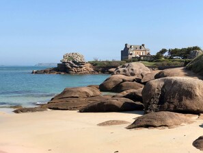 A coastal landscape with large boulders, a sandy beach, and a historic house with a garden.