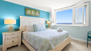 A bedroom with a bed, nightstands, a lamp, a sign reading "SEA", and a window with a view of the ocean. - 🌊 Views That Go On Forever (Destin)