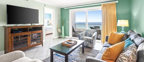 A living room with a flat-screen TV, a wooden entertainment unit, a sofa, and a coffee table. There is a balcony with a view of the ocean.
