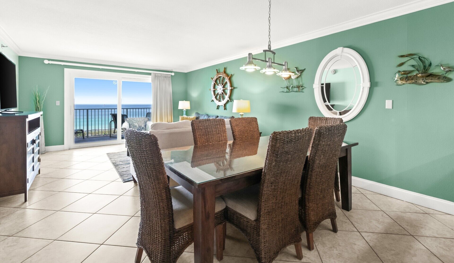 A dining area with a glass table and wicker chairs, a chandelier, a sail wheel decoration, and a view of the ocean through a sliding door.