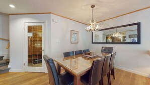 A dining room with a glass-top table, six chairs, a chandelier, a large mirror, and a wooden door leading to a kitchen.