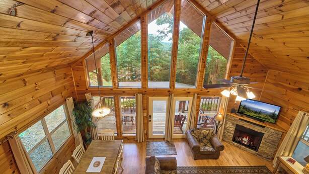 Eine Hütte mit hohen Decken, Holzwänden, einem Kamin, einem Fernseher und einem großen Fenster mit Blick auf einen Wald.