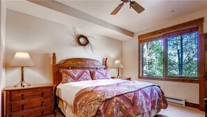 A bedroom with a wooden bed, two bedside lamps, a wooden nightstand, a decorative wreath, and a large window with a view of trees. - Tl2212 Stunning Condo with Private Balcony Overlooking the Pool (Steamboat Springs)