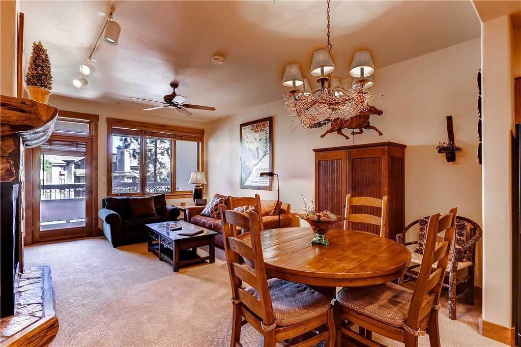 Un vaste salon doté d’une table à manger ronde en bois, de sièges confortables, d’un ventilateur de plafond et de grandes fenêtres offrant une vue dégagée.