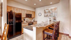A kitchen with wooden cabinets, a black refrigerator, a microwave, and a dining area with a bar stool. - Tl2212 Stunning Condo with Private Balcony Overlooking the Pool (Steamboat Springs)
