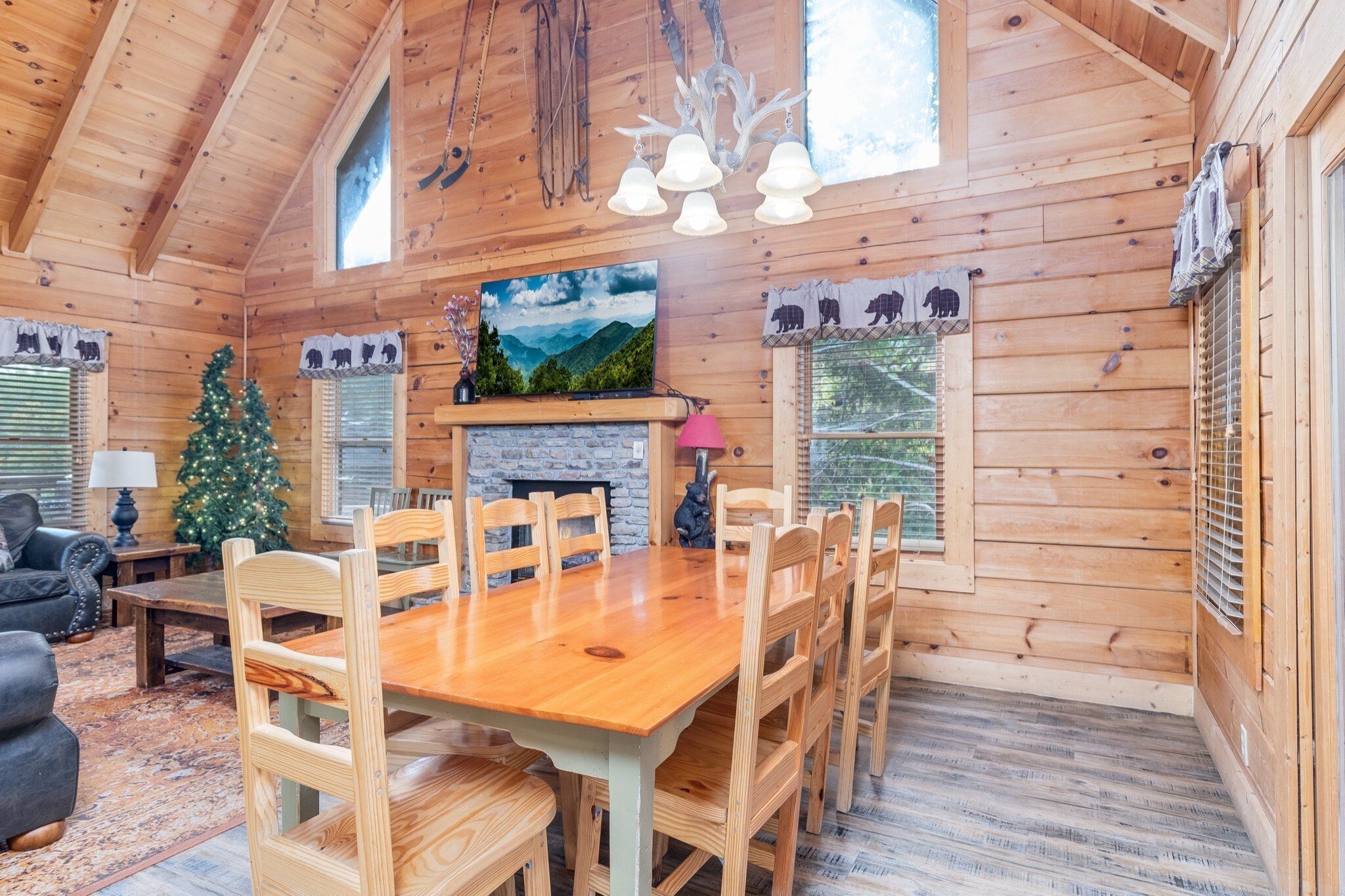 A wooden cabin interior with a dining area, fireplace, Christmas tree, and a painting of a mountain landscape.