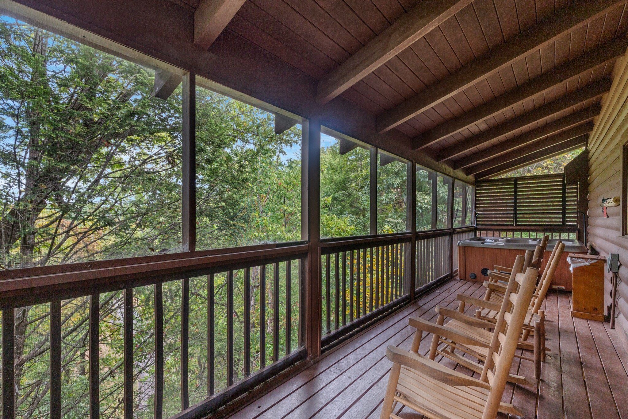 A wooden deck with a view of trees, a hot tub, and wooden chairs.