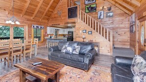 A wooden cabin interior with a black leather sofa, wooden dining table, and a staircase leading to a loft area. - ER101 - Heavenly Hideaway  Great location - Close to town! (Pigeon Forge)