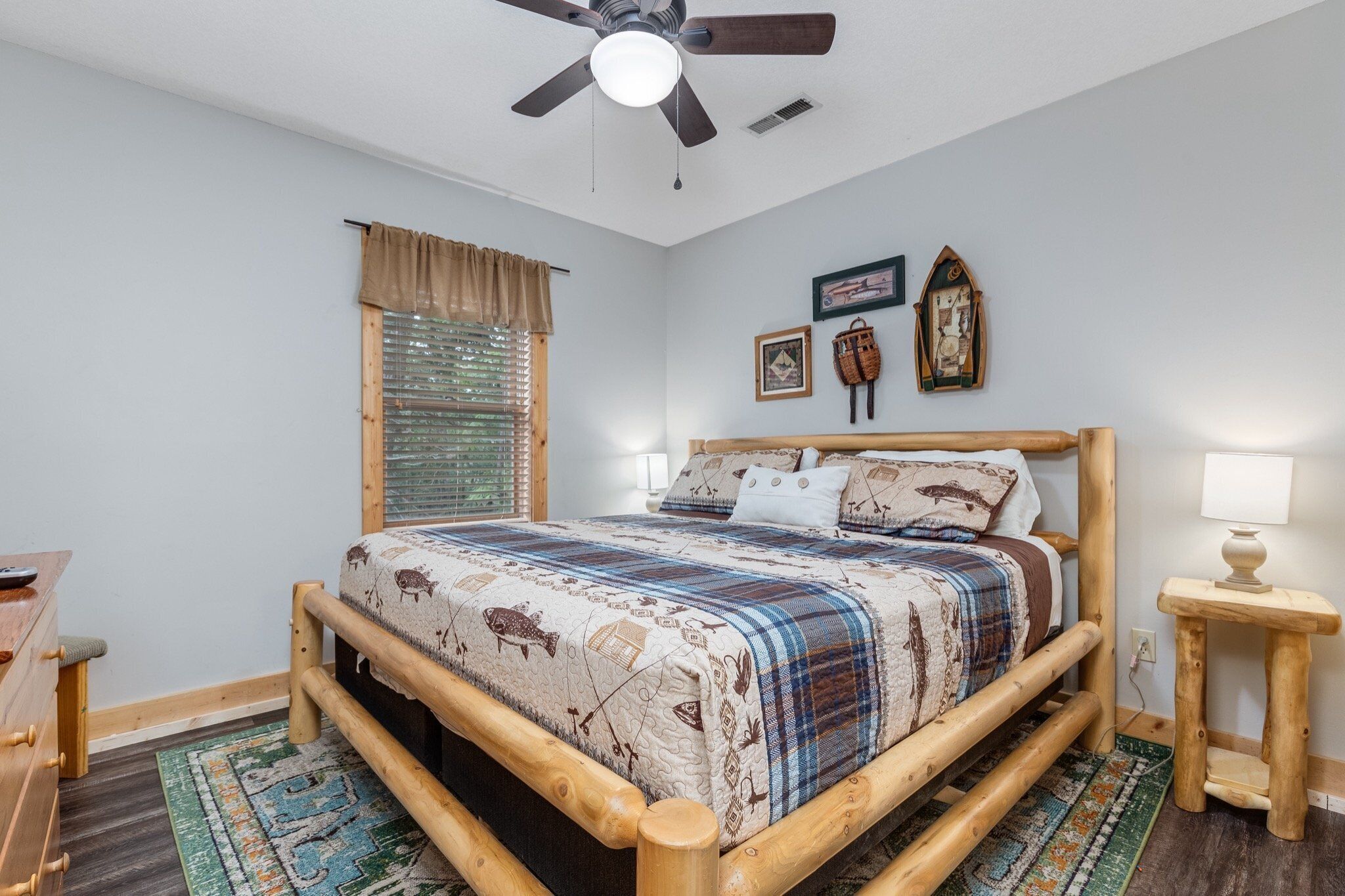 A cozy bedroom with a wooden bed frame, a ceiling fan, a window with blinds, and a bedside table with a lamp.
