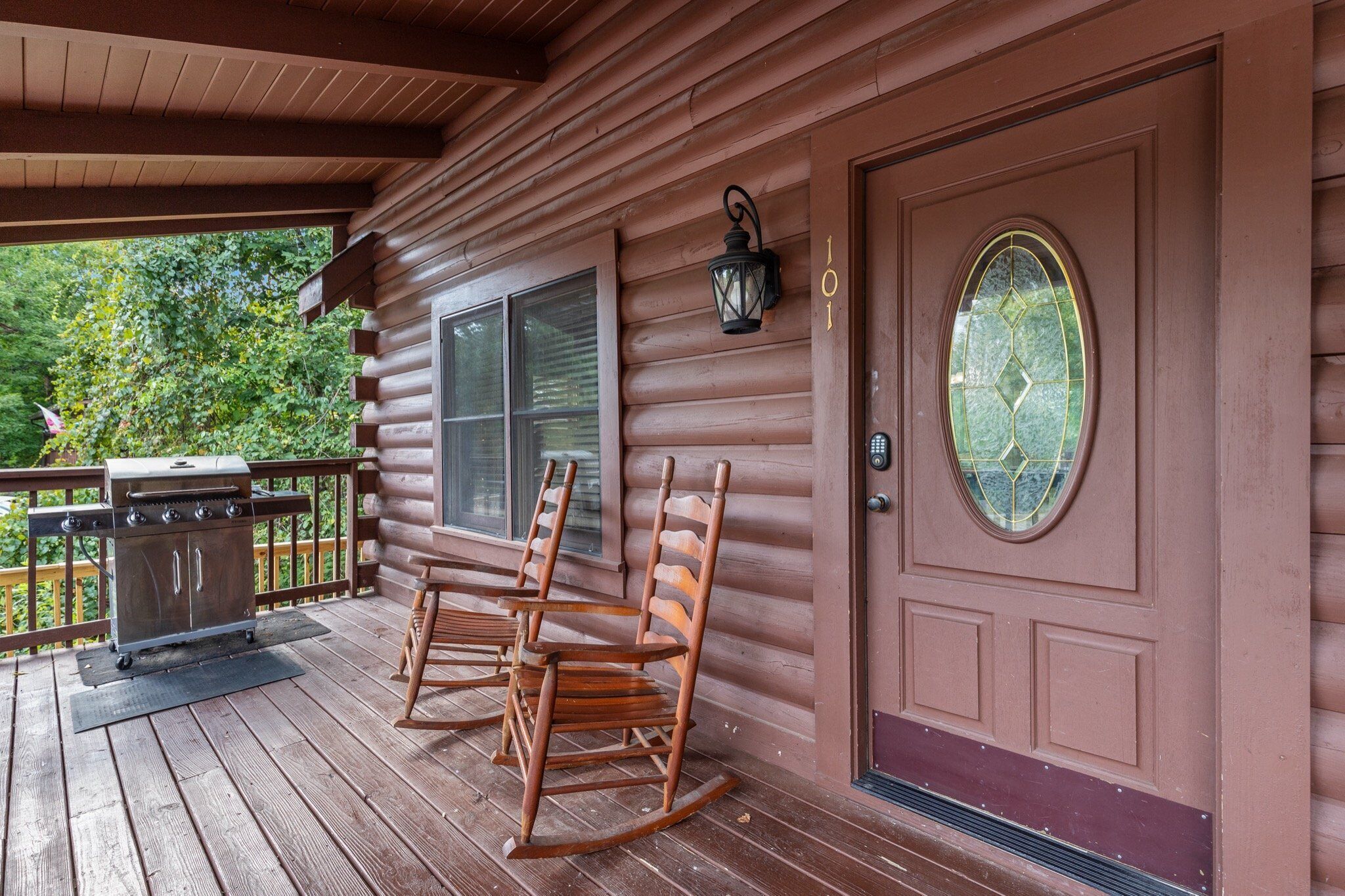 A wooden porch with a grill, rocking chairs, and a door with a glass panel.