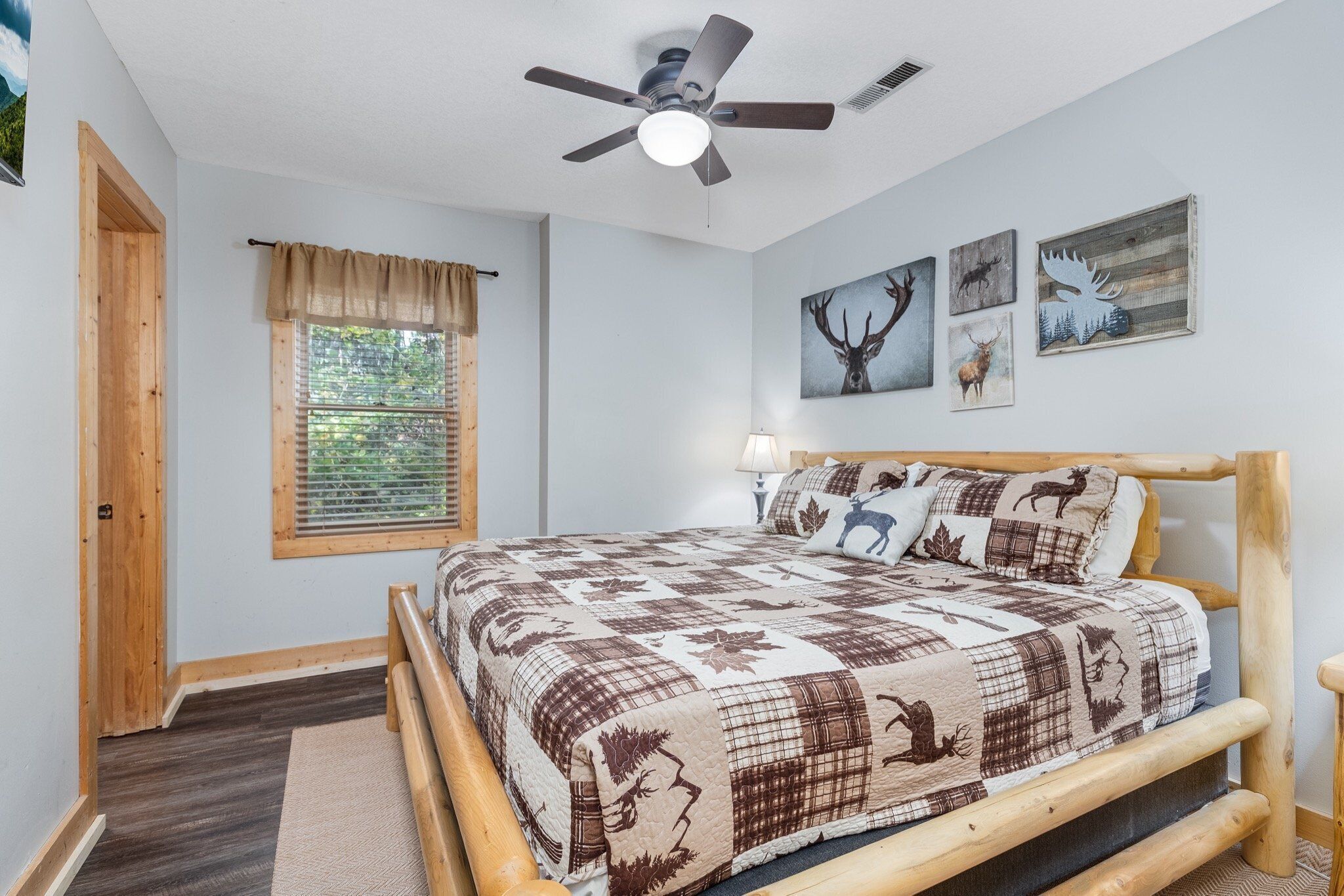 Une chambre à coucher avec un lit en bois, un ventilateur de plafond, une fenêtre avec des stores et des œuvres d’art murales représentant des cerfs et des élans.