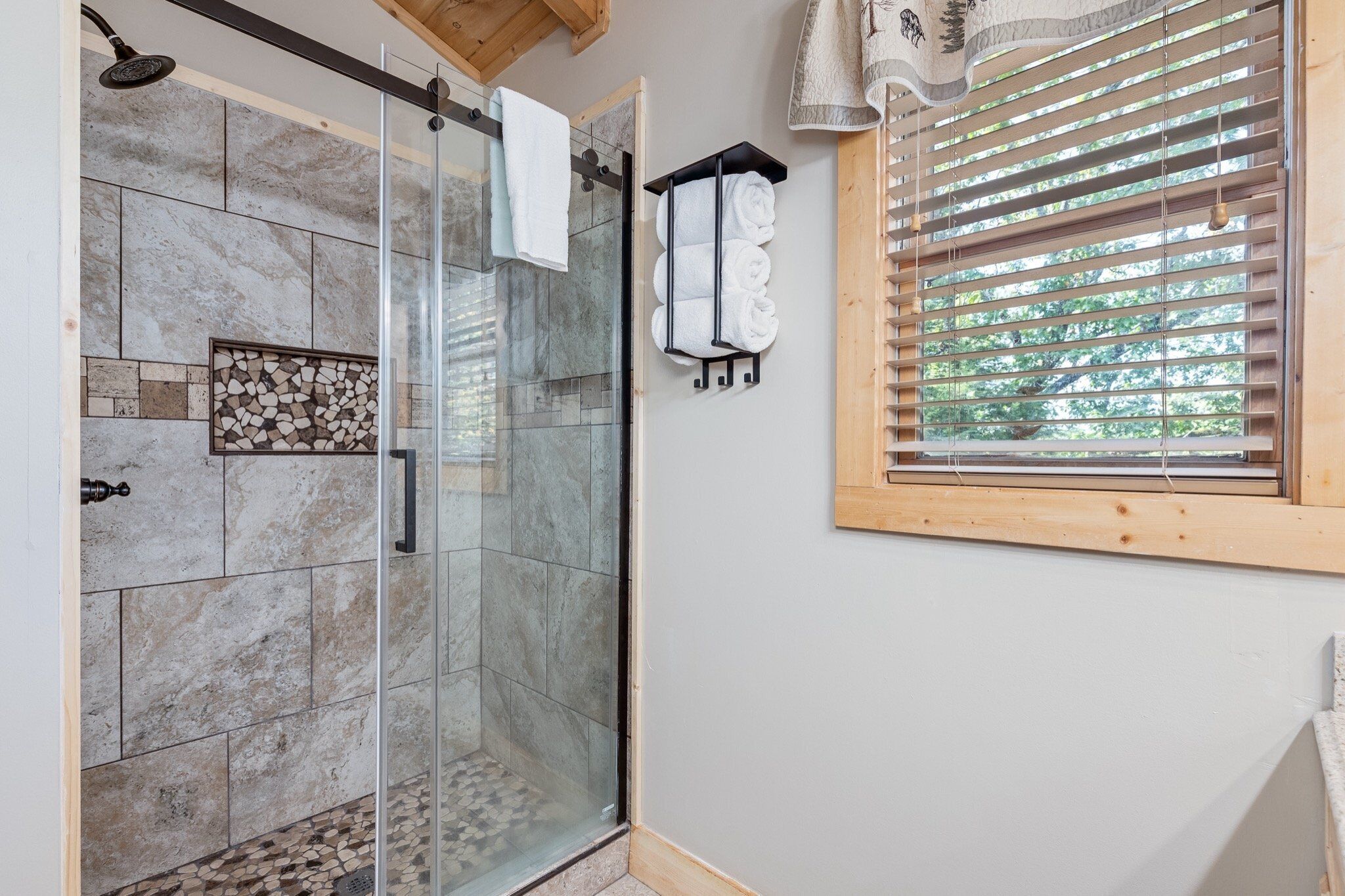 Une douche fermée par des parois de verre, avec un mur carrelé, un plafond en bois et une fenêtre équipée de stores.