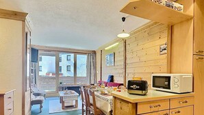 A compact kitchen with wooden cabinets, a microwave, and a toaster. There is a dining area with a table and chairs, and a view of the outside through a window.