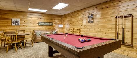 A pool table with surrounding chairs and a table, wooden walls, and a billiards rack.