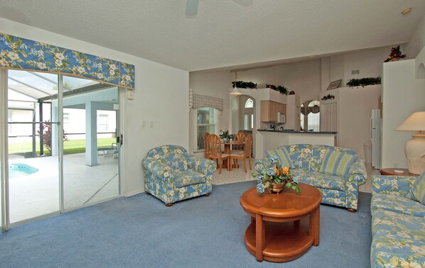 A living room with a blue carpet, two floral-patterned sofas, a coffee table, and a glass door leading to a patio with a pool.