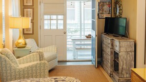 Une pièce avec une porte blanche donnant sur un balcon, un téléviseur posé sur une commode en bois, des chaises à motifs et une lampe sur une table d’appoint.