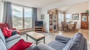 A spacious living room with a large window, a television, a bookshelf, and a dining area with a table and chairs.