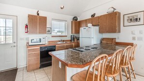 A compact kitchen with wooden cabinets, a built-in microwave, a sink, a stove, and a refrigerator. There are three wooden chairs around the island.