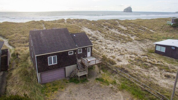 A two-story house with a dark roof and a front porch, surrounded by dunes and a beach.