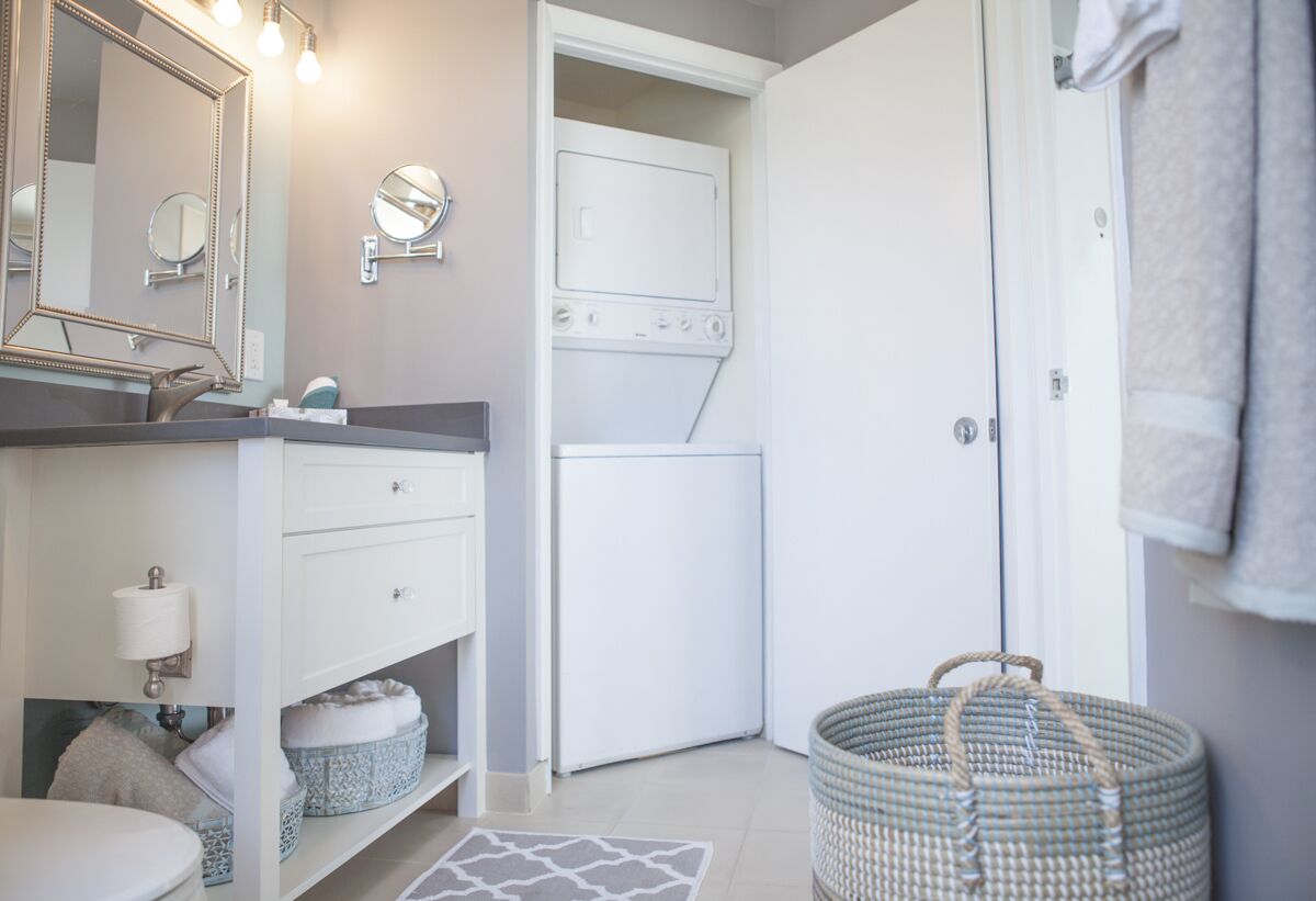A modern bathroom with a large mirror, a sink, a toilet, a washing machine, and a basket.