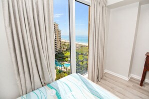 Une chambre d’hôtel avec une grande fenêtre, un lit recouvert d’une courtepointe à rayures et une vue sur un complexe hôtelier doté d’une piscine et d’une plage.