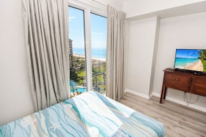 A bedroom with a large window, a bed with a striped bedspread, a wooden nightstand, and a television displaying a beach scene.