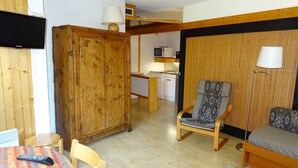 Um espaço compacto com guarda-roupa de madeira, uma mesa de jantar com cadeiras, um sofá, uma poltrona estampada, uma pequena cozinha e uma televisão.