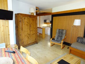 Un espacio habitable compacto con un armario de madera, una mesa de comedor con sillas, un sofá, un sillón estampado, una pequeña zona de cocina y un televisor.