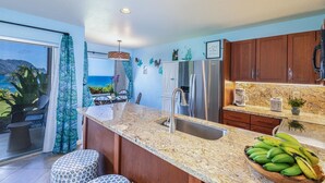 A kitchen with a view of the ocean, a refrigerator, a sink, and a countertop with bananas.