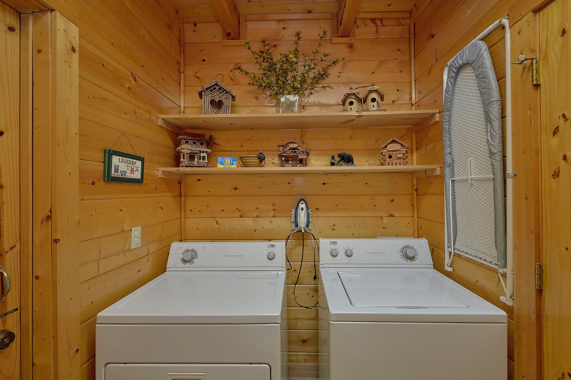 A laundry room with two white washers and dryers, wooden walls, and shelves with decorative items.