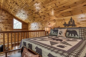 Ein Schlafzimmer in einer Blockhütte mit einem Bett, einem Fenster, einem Nachttisch und Blick auf Bäume.