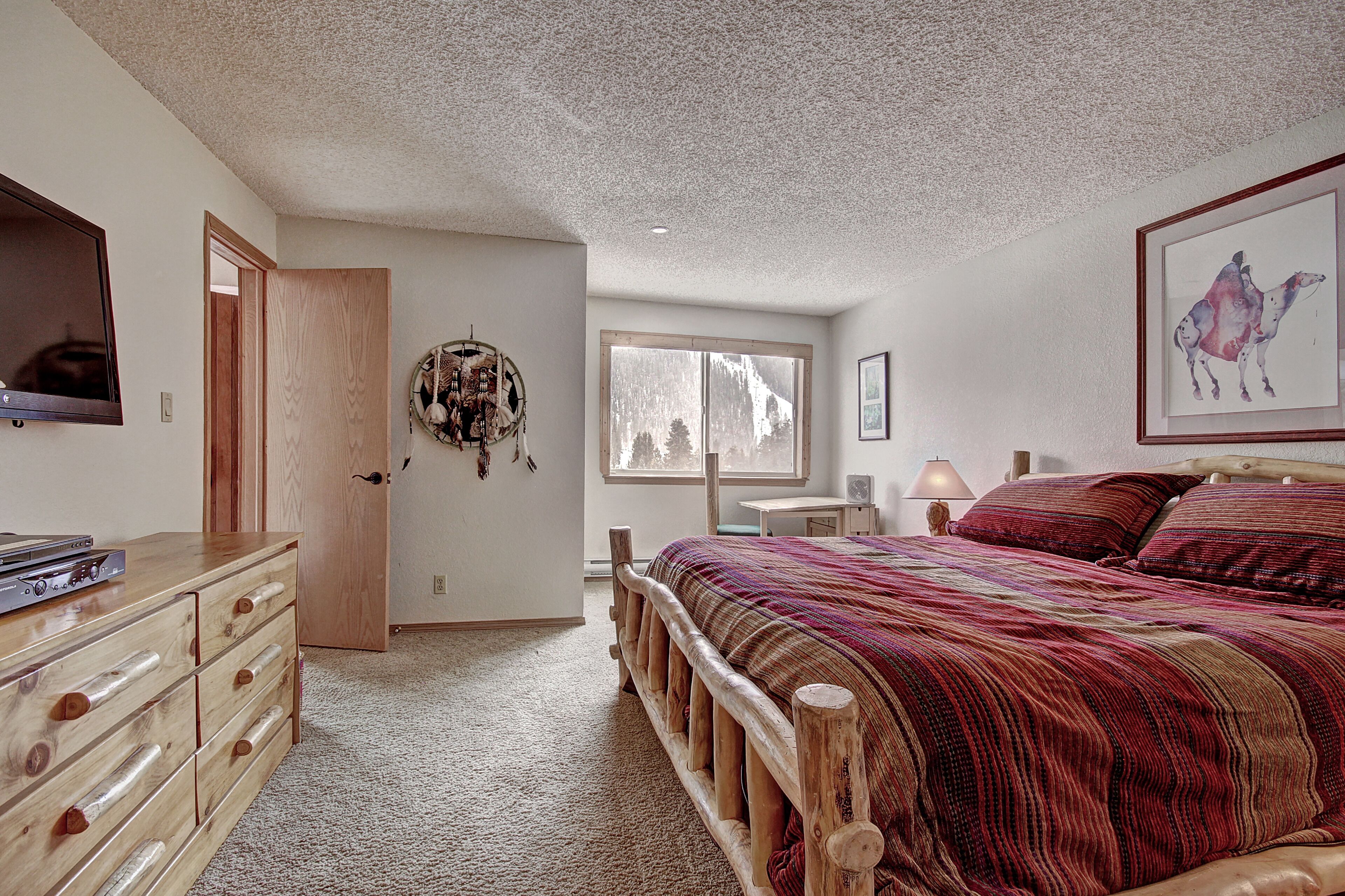 A bedroom with a wooden bed, a dresser, a mirror, a window, and a wall-mounted artwork.