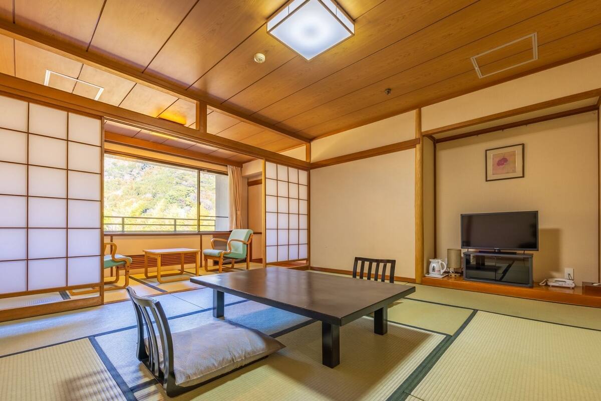 Una habitación japonesa tradicional con suelo de tatami, una mesa baja, sillas, un televisor y una ventana con vistas a la vegetación.