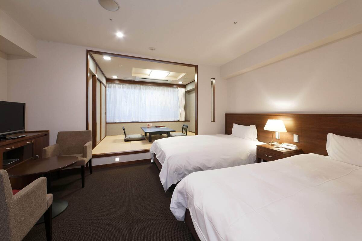 Habitación de hotel con dos camas, un escritorio, una silla, un televisor y una ventana con cortinas.