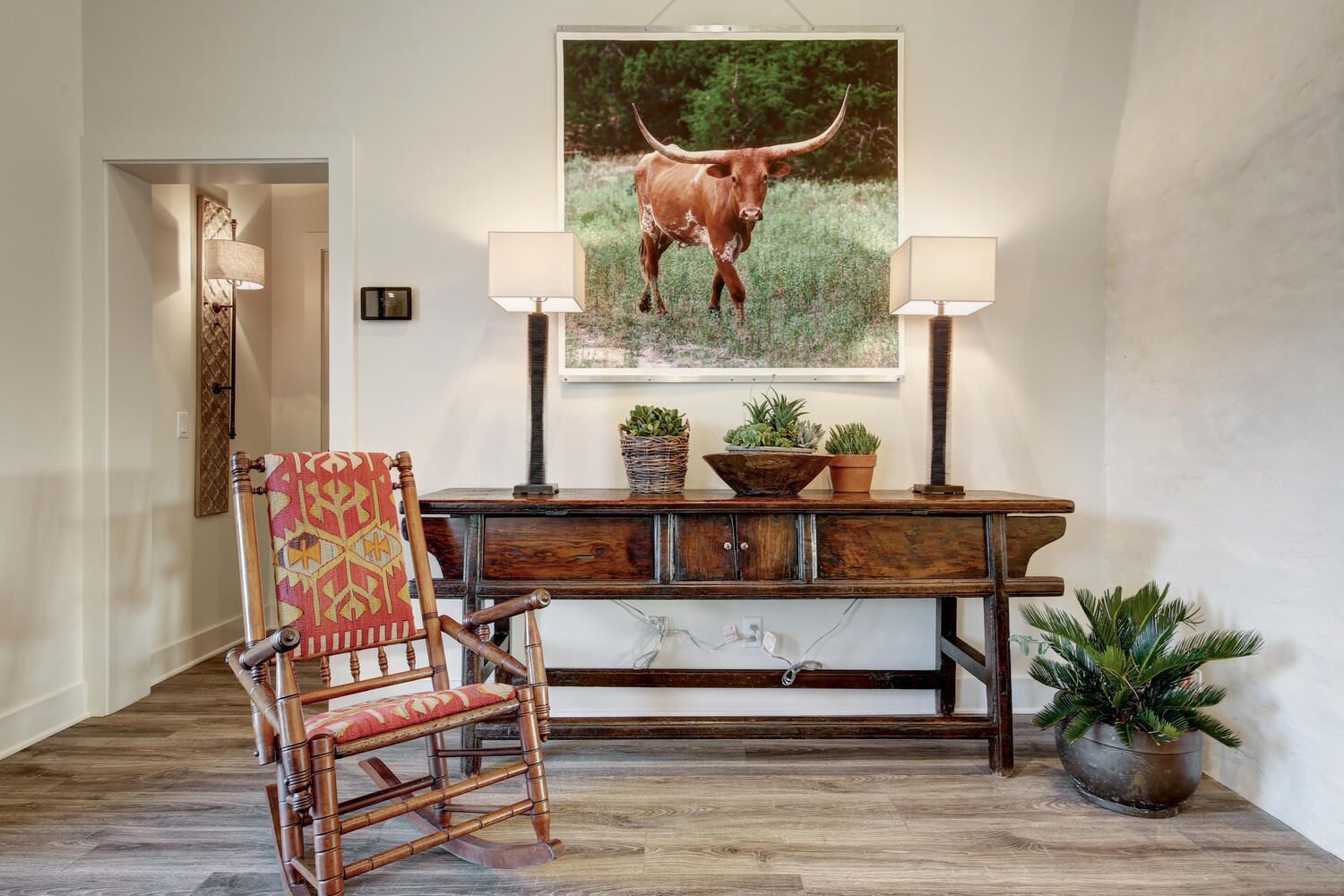 Une chaise en bois avec un coussin à motifs, une console en bois ornée de plantes en pot et, au mur, un tableau encadré représentant un taureau.