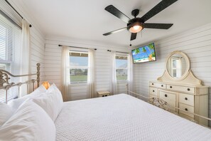 Une chambre à coucher avec un lit, un ventilateur de plafond, une commode, un téléviseur et une fenêtre avec des rideaux.