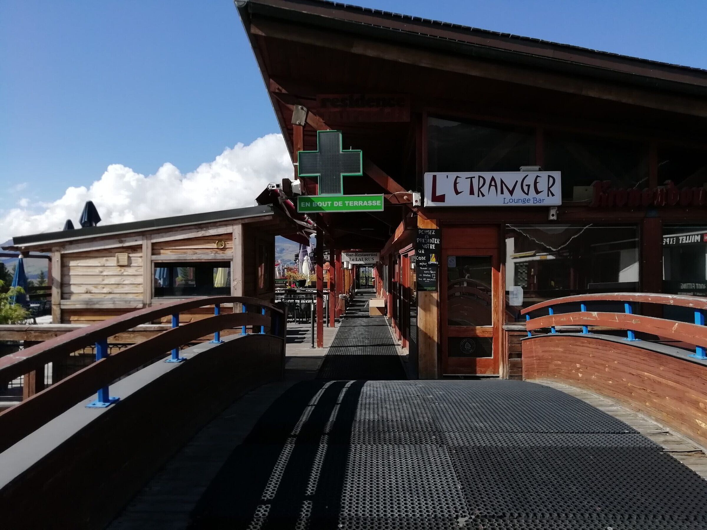 Banchina di una stazione ferroviaria con una struttura in legno, un cartello che indica 'L'etranger Lounge Bar' e un segnale verde con scritto 'EN BOUT DE TERRASSE'.