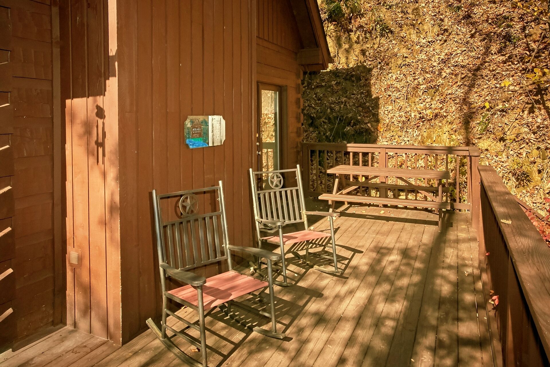 Een houten terras met metalen schommelstoelen en een picknicktafel, omgeven door een stenen muur en een houten gebouw.