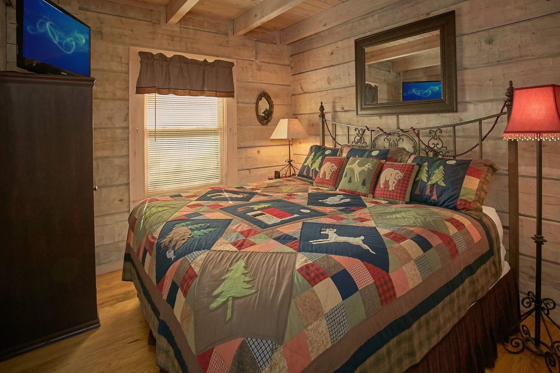 A bedroom with a wooden floor, a bed with a colorful quilt, a bedside lamp, a television on a stand, and a window with blinds.
