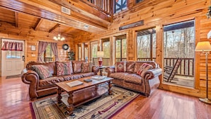 Ein Interieur einer Hütte mit Holzboden, Ledersofas, einem Couchtisch und großen Fenstern mit Blick ins Freie.