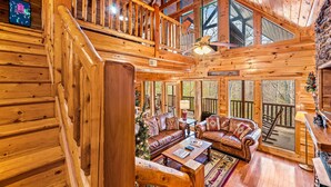 Ein großzügiges Blockhaus-Interieur mit einer zentralen Sitzecke, die ein Sofa und einen Couchtisch umfasst, einer Treppe zu einer oberen Ebene sowie großen Fenstern mit Blick auf den umliegenden Wald.