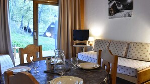 Une pièce chaleureuse avec un plafond en bois, un canapé, une télévision, une table à manger dressée avec des assiettes et des verres, et une vue sur les arbres à l’extérieur.
