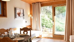 Un coin repas avec une table dressée pour deux personnes, un plafond en bois et une vue sur un jardin, visible à travers une porte vitrée.