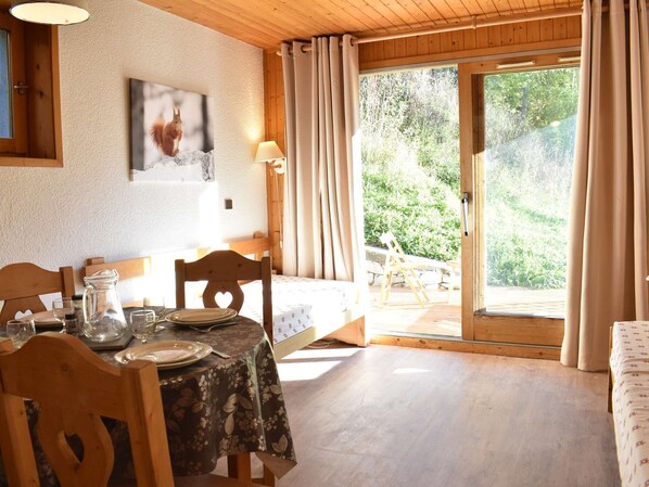 A dining area with a table set for two, a wooden ceiling, and a view of a garden through a glass door. - Charming Studio for 4 in Méribel, Near Slopes & Shops, Balcony with Mountain Views (Méribel)