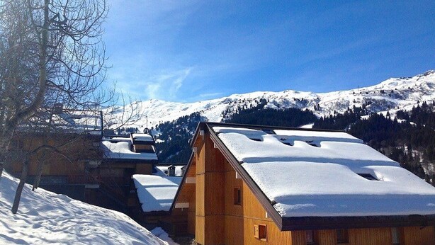 Une cabane en bois, dont le toit est recouvert de neige, se dressant sur fond de montagnes enneigées et d’un ciel bleu clair.