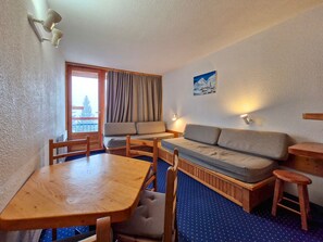 Een kamer met een houten tafel, twee stoelen, een bank, een klein krukje en een raam met gordijnen.