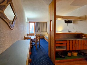 A compact room with a dining table, chairs, a sofa, a small kitchenette, and a window with curtains.